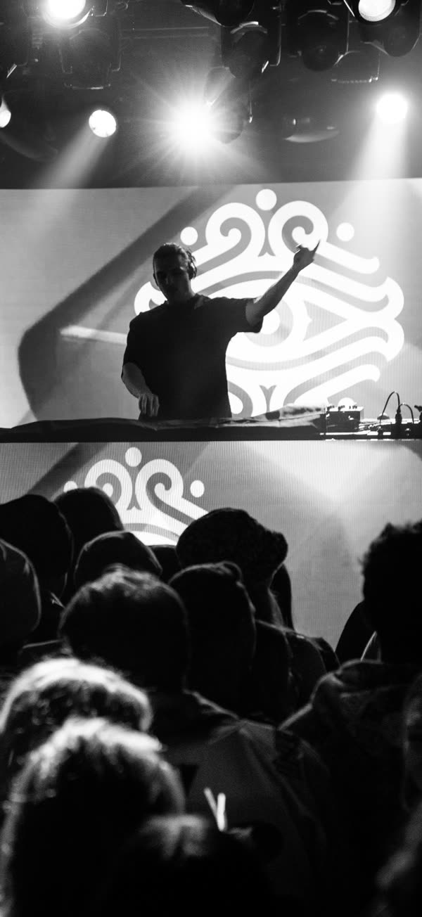 Alec Hollingsworth behind the decks at a live set, the illoh mark on the screen behind him